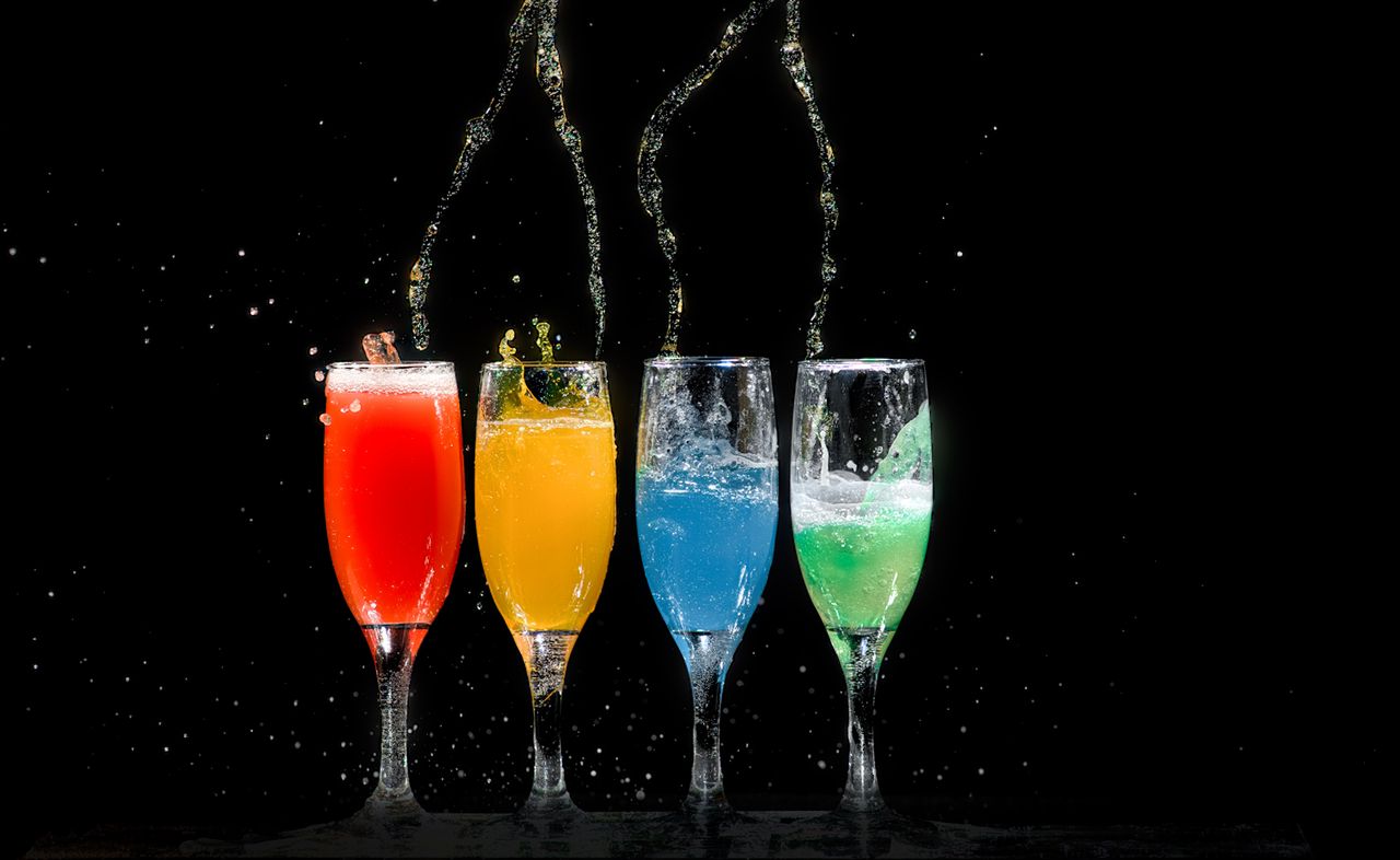 Vibrant colored cocktails splashing in glasses against a black background.